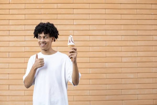 糖果和人们的观念 微笑着 卷曲头发的非裔美国人 拿着一个冰激淋筒 竖起大拇指 把目光移开 穿着一件白色T恤放在砖墙上 — 图库照片
