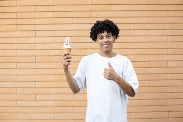 糖果和人们的观念 一个快乐的 卷曲头发的非裔美国人 手里拿着一个冰激凌筒 竖起大拇指 穿着一件白色T恤 站在砖墙的背景上 — 图库照片