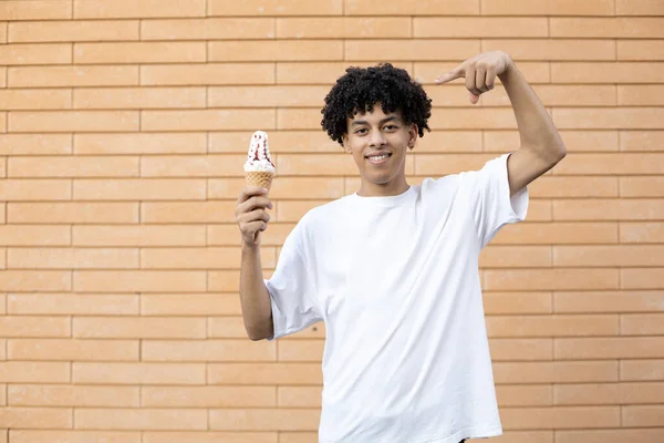 垃圾食品 糖果和人的概念 满意的卷曲的非裔美国人 手里拿着冰淇淋 从上面指着冰淇淋 穿着一件白色T恤衫在砖墙上 — 图库照片
