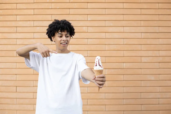 人们的概念 美国人把一个白色的冰淇淋锥放在伸出的手上 用手指指着它 穿着一件白色的T恤 贴在砖墙的背景上 上面有复制的空间 — 图库照片