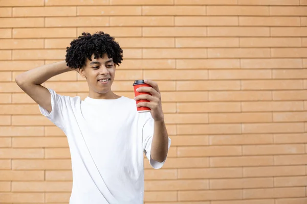 生活方式和人们的观念 快乐的卷曲的非裔美国人 手里拿着一杯红葡萄酒咖啡 手背在身后 穿着一件白色的T恤 在砖墙上看着它 — 图库照片