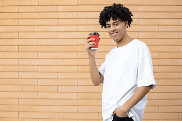 生活方式和人们的观念 笑着英俊的非洲裔美国男人 手里拿着一杯红咖啡 穿着一件白色T恤 站在砖墙的背景上看着摄像机 — 图库照片