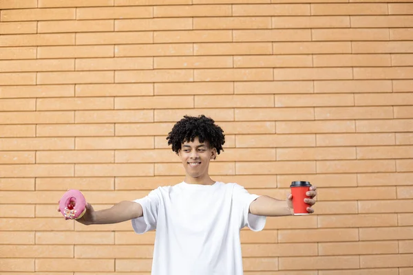 生活方式和人的观念 快乐英俊的非裔美国男人 拿着一杯红咖啡和一个粉色的甜甜圈 看着它 穿着外面砖墙上的一件白色T恤 — 图库照片