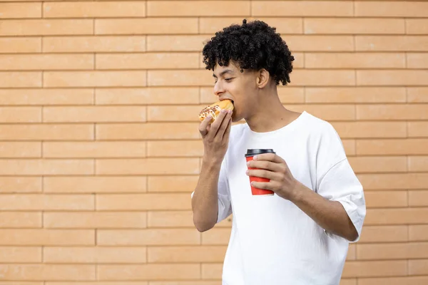 生活方式和人的概念 微笑着拿着一杯咖啡咬着橙子甜甜圈的非裔美国人 凝视着砖墙背景上的一件白色T恤 — 图库照片