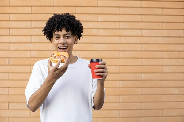 デザート ドリンク ライフスタイル 人々のコンセプト 幸せなアフリカ系アメリカ人男性がオレンジのドーナツをかむとコーヒーのカップを保持し カメラを見て レンガの壁に白いTシャツを着て — ストック写真