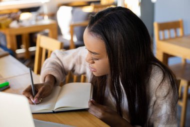 İş, teknoloji, yaşam tarzı ve insan kavramı. Bilgisayarlı girişimci bir kız, bir kafede çevrimiçi bir konferans sırasında günlüğüne önemli notlar yazıyor.
