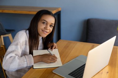 Çevrimiçi eğitim, teknoloji, yaşam tarzı ve insan konsepti - dizüstü bilgisayarlı gülümseyen kız öğrenci, defterine kalemle notlar yazması, kahve dükkanındaki kameraya bakması