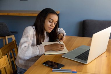 Teknoloji, yaşam tarzı ve insan konsepti. Elinde not defteriyle gülümseyen siyah saçlı Afro-Amerikalı bir kız. Bir kafedeki online toplantı sırasında not defterine kalemle notlar yazıyor.