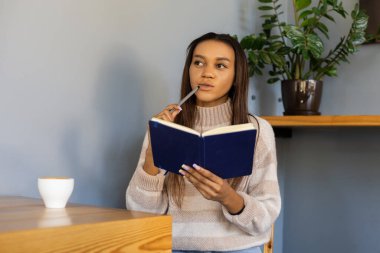 Çevrimiçi eğitim, teknoloji, yaşam tarzı ve insan konsepti - Bir not defteri ve bir fincan kahveyle düşünceli bir kız, yüzüne kalem tutarak, bir salgın sırasında oturup ders çalışarak