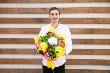 Beyaz tişörtlü gülümseyen beyaz adam elinde bir buket çiçekle kahverengi kurdeleli arka planda fotokopi çekiyordu.
