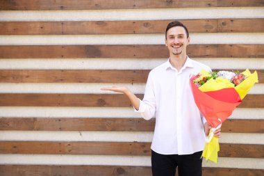 Jest, çiçekler, duygular ve insan konsepti - beyaz gömlekli ve siyah kotlu gülümseyen Avrupalı adam büyük bir buket tutuyor ve elini kahverengi kaburgalı arka plandaki boşluğu kopyalamak için işaret ediyor