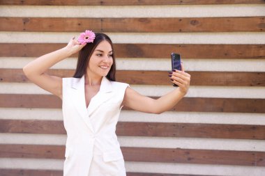 Beyaz elbiseli, pembe çiçekli, gülümseyen doğulu bir kız telefonunda fotokopi alanı olan kahverengi kurdeleli bir fonda selfie çekiyor.