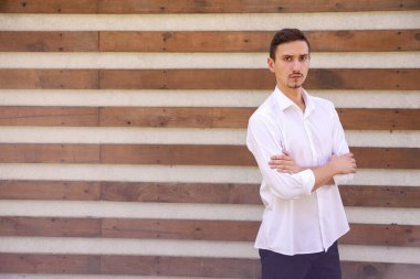 Beyaz gömlekli ciddi sakallı bir Avrupalı. Kolları çapraz ve fotokopi çekilen kahverengi bir arka planda kameraya bakıyor.