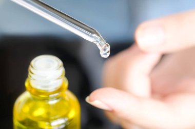 Güzel bir kadının elleri, yüzüne serum kolajen nemlendirici dökerek berrak sarı şişe yumuşak ışık arka planına döküyor. Koruyucu cilt bakımı yüz özü yağı, C vitamini güzellik ve kaplıca konsepti..