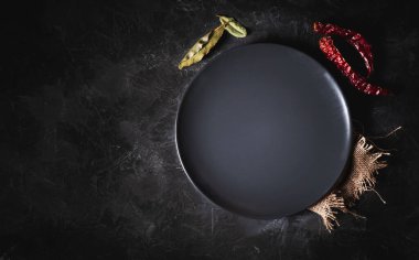 Karanlık bir masanın üzerine boş yuvarlak siyah tabak ve yemek pişirmek için bazı malzemeler. Kurutulmuş biber ve defne yaprağı.