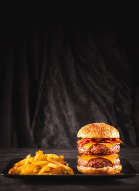 Duble sığır eti ve pastırmalı burger, çedar peyniri ve siyah bir tabakta patates cipsi, yenmeye hazır. Fotoğraf alanı kopyala. 