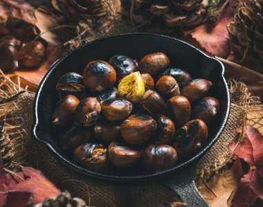 Sonbahar yaprakları ve kozalaklarla dolu bir masada küçük demir bir tavada kavrulmuş kestane. Yemek hazır. Geleneksel sonbahar yemekleri.