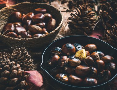 Çiğ ve kavrulmuş kestaneler küçük bir demir tavada ve sonbahar yaprakları ve çam kozalaklarıyla dolu bir masada bir kase. Yemek hazır. Geleneksel sonbahar yemekleri.