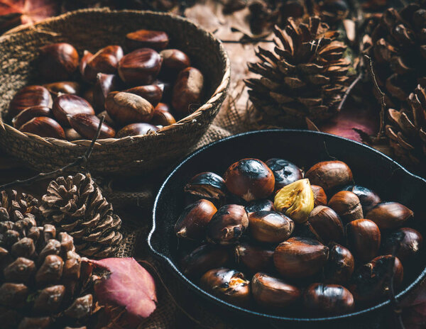 Raw and roasted chestnuts in a small iron pan and a bowl on a table with autumn leaves and pine cones. Ready to eat. Traditional autumn food.
