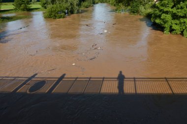 Köprüde duran bir adamın silueti. Dalgalar kahverengi su kütleleri tarafından sürüklenir. Arka planda ağaçlar var. Almanya 'da sel. Yüksek açı görünümü.