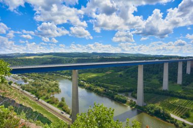 Mavi çelik ve gri sütunlardan yapılmış güzel Mosel Vadisi köprüsü, arka planda büyük ağaçlar, beyaz gri bulutlu mavi gökyüzü, demiryolu rayları görülebilir. Almanya.