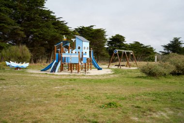 Ormanın kenarındaki oyun parkında. Kumlu zeminde slaytlar ve salıncaklarla mavi oyun binası. Fransa, Britanya.