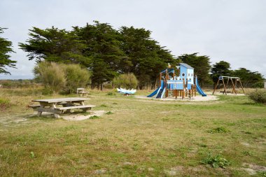 Ormanın kenarındaki oyun parkında. Kumlu zeminde slaytlar ve salıncaklarla mavi oyun binası. Önünde masa olan banklar. Fransa, Britanya.