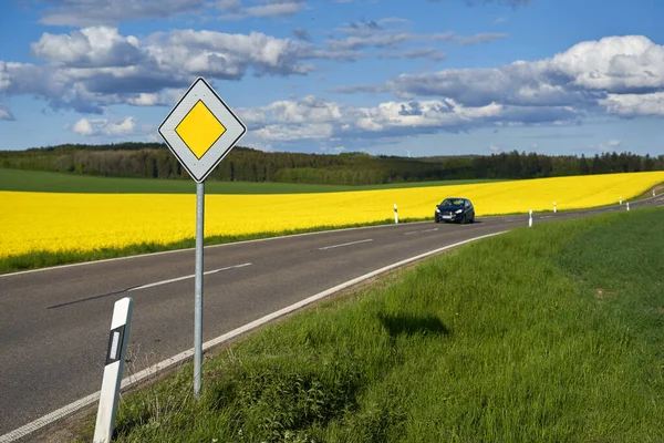 Traffic sign on the side of the road. Yellow rapeseed (Brassica napus) field behind the roadway. 1 Car driving on the track. Green forest in front of blue cloudy sky.