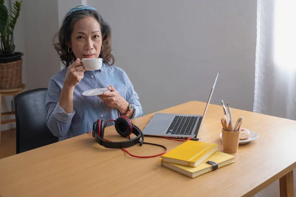 relax time, Portrait of an older woman sitting on her laptop listening to music and having a drink to relax inside the house.