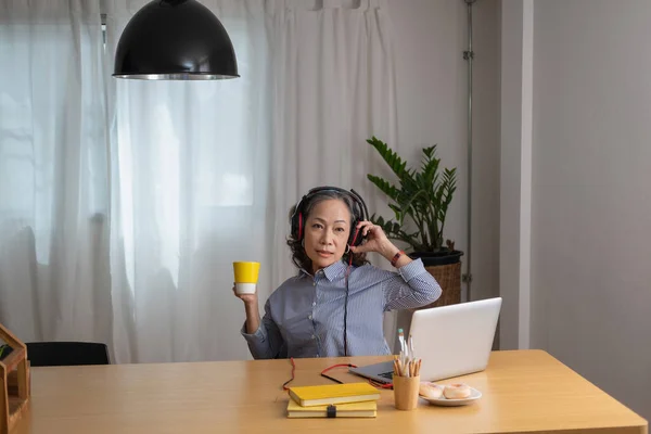 relax time, Portrait of an older woman sitting on her laptop listening to music and having a drink to relax inside the house.