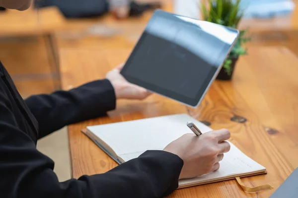 A businesswoman holds a pen to write a message on a notebook and holds a tablet to record company information.