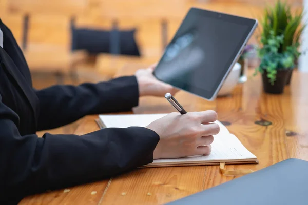 A businesswoman holds a pen to write a message on a notebook and holds a tablet to record company information.