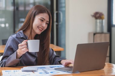 İş kadını, dizüstü bilgisayarını ve belgelerini kullanarak şirket satışlarını analiz etmeden önce elinde bir kahve fincanı tutuyor..