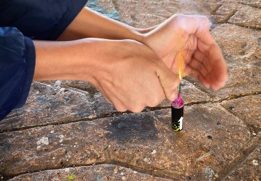 hands of a naughty child with a lighter lighting a large firecracker on the ground, sparks flying from the wick of the explosive about to go off and explode, horizontal