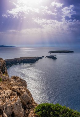 Günbatımından saatler önce kayalık uçurumlardan deniz manzarası, küçük bulutların arasından süzülen güneş ışınları, dikey, Fornells, Menorca, Balearic Adaları, İspanya