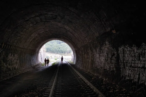 the exit of an old dark railroad tunnel seen from the inside with the silhouette of three people in the distance coming out of the tunnel, concept of overcoming
