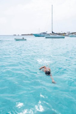 a boy diving near some sailboats and yachts in a cove of crystal clear turquoise water, clear sea, boy in foreground swimming with a snorkel, copy space, in Menorca, Balearic Islands, Spain