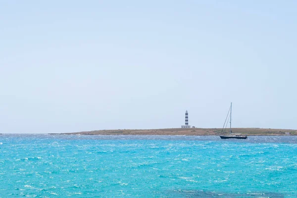 a sailboat skirts a cape with a blue and white lighthouse, on a sea of turquoise waters, clear sky, Isla del Aire, Punta Prima, Menorca, Spain, copy space