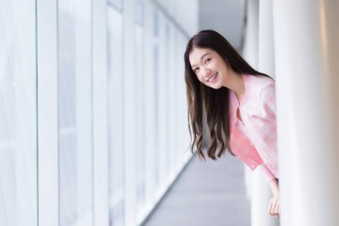 Güzel genç Asyalı kız pembe gömlekli beyaz bir elbise giyer ve güzel bir günde cam pencerenin yanında dikilirken neşeyle gülümser..