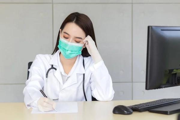 アジア系の女医は患者の治療記録を書きながら真剣に働く 彼女は医療コンセプトで病院の臨床検査室で医療マスクとラテックス手袋を着用しています — ストック写真