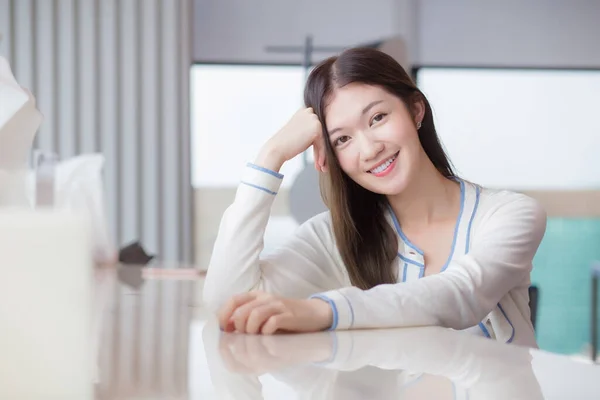 Beautiful adult young age Asian professional woman with long hair in white shirt looks at the camera her smiling happily while she sits on chair in workplace office with glassed building as a background.