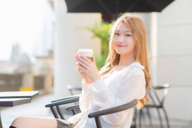 Bronz saçlı Asyalı güzel bayan kahve dükkanında sandalyeye oturup gülümserken güneşli bir sabahta elinde kahve fincanı tutuyor..