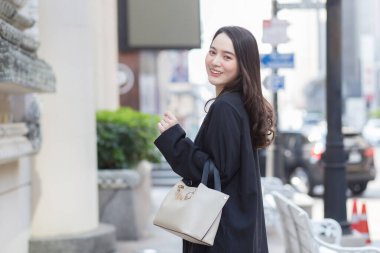 Siyah ceketli, güzel, uzun saçlı bir Asyalı kadının portresi, yürüyüş çantası taşıyor, açık havada mutlu bir şekilde gülümsüyor..