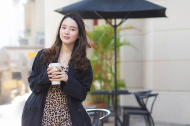 Koyu mavi takım elbiseli genç Asyalı güzel bir kadın güneşli bir sabah kahve dükkanının önünde elinde mutlu bir kahve fincanı tutuyor.