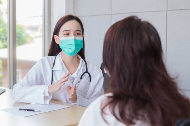 Genç Asyalı profesyonel kadın doktor, yaşlı bir kadın hastaya semptomlarını dikkatlice incelerken, ikisi de sağlık hizmetleri konseptindeki hastane muayene odasında enfeksiyonu önlemek için maske takıyor..