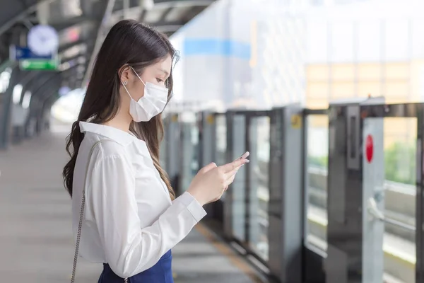 年轻的亚洲女商人戴着面具站在城中的天空火车站上 一边用她的智能手机向顾客办公室传递信息 — 图库照片