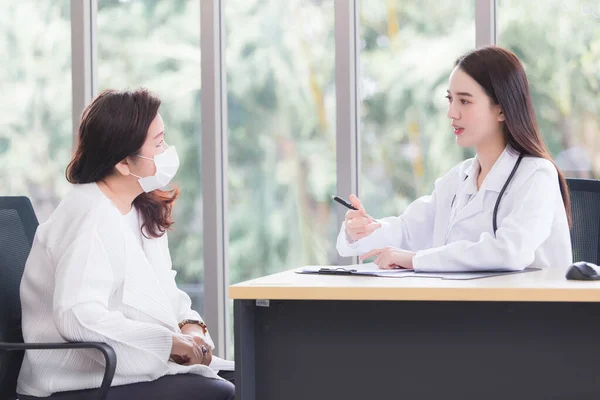 Asian woman doctor is giving advice on symptoms or health problems to an elderly female patient in an examination room at a hospital.