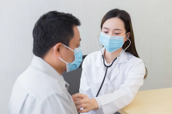 アジアの男性患者は健康診断を受け 女性医師は聴診器を使用してコロナウイルスパンデミックの心拍数を聞く病院の診察室で外科用マスクを常に着用することによって コロナウイルス保護の概念 — ストック写真