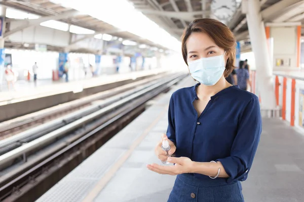 亚洲妇女正在城市的户外车站等火车 当她看着相机时 她喷了些酒精来清洁她的手 并戴着医疗面罩作为医疗保健和新的正常生活方式 — 图库照片