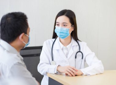 Profesyonel Asyalı kadın doktor, Coronavirus hastalığını önlemek için hastanedeki muayene odasında yüz maskesi takarken bir hastayla ağrısı ve semptomları hakkında konuşuyor. Sağlık ve çevre kirliliği